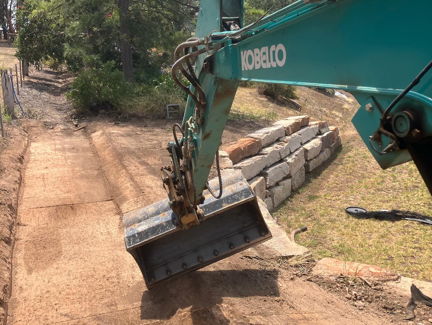 Branded Kobelco excavator mid-build at a rural stone retaining wall